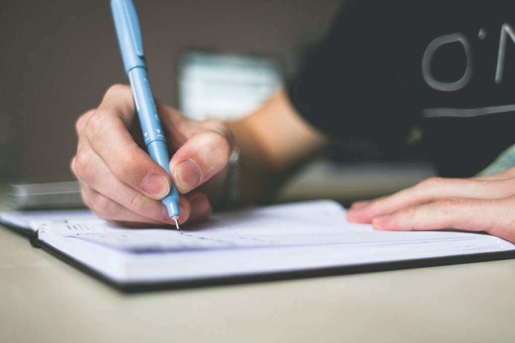 The image shows someone holding a pen and writing in a notebook at a table. The focus is on the person's hand, and their body is blurred in the background. 